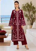 A woman models a deep maroon three-piece kurta set with intricate white floral embroidery, standing in a bright, modern living room with a view of the ocean in the background.