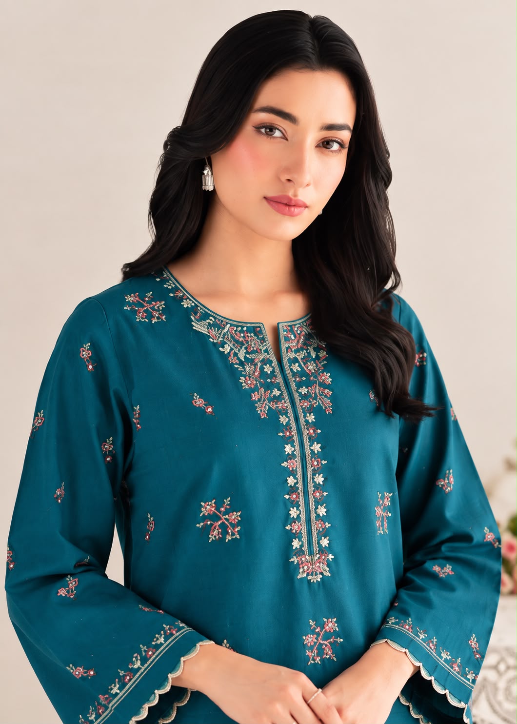 A woman wearing a teal blue embroidered kurta with floral patterns and scalloped cuffs, photographed against a soft beige background.