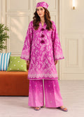 A woman modeling a bright pink traditional outfit with a matching headscarf, featuring a floral patterned kurta with decorative buttons and wide-leg trousers, set against an indoor background with a sofa and cushions.