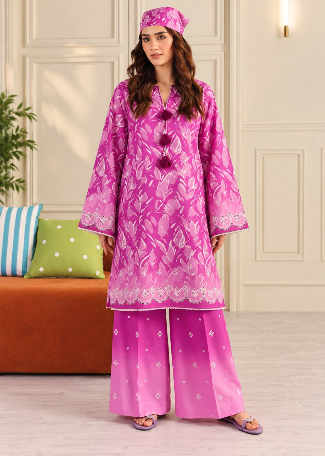A woman modeling a bright pink traditional outfit with a matching headscarf, featuring a floral patterned kurta with decorative buttons and wide-leg trousers, set against an indoor background with a sofa and cushions.