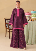 A woman modeling a traditional Indian outfit consisting of a purple kurta with pink embroidery and matching wide-leg trousers with a floral print, set against a warm beige background with a decorated table and chair.