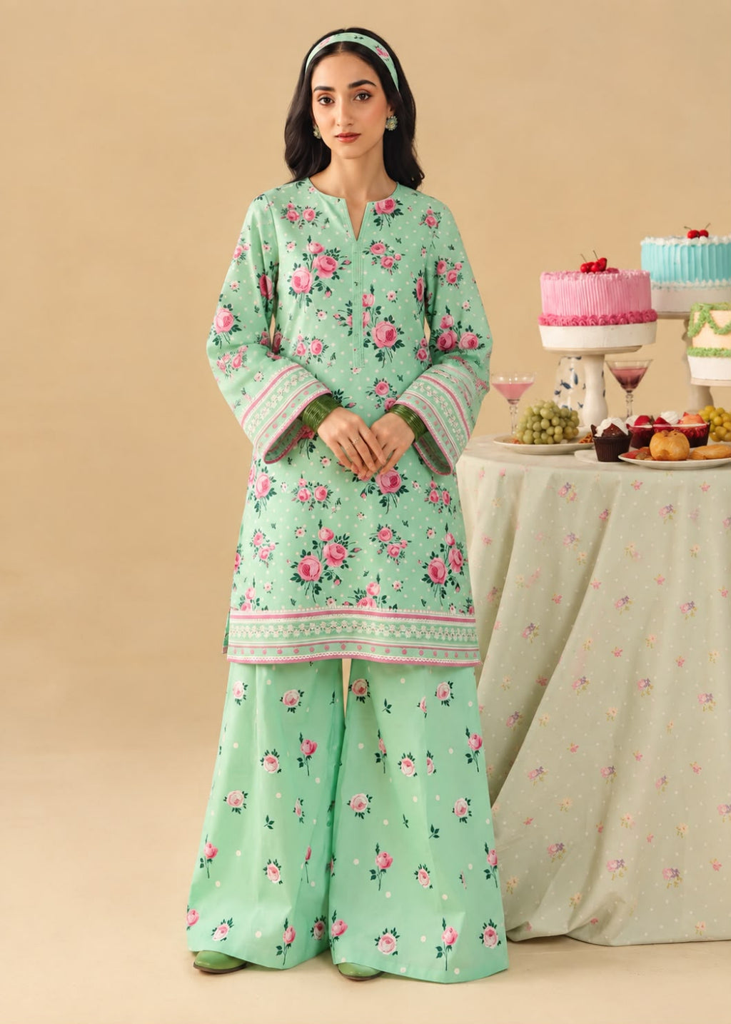 A woman models a mint green three-piece traditional outfit with pink floral prints and green borders, standing in front of a beige backdrop next to a decorated table.