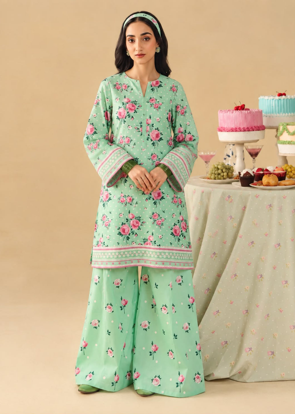A woman models a mint green three-piece traditional outfit with pink floral prints and green borders, standing in front of a beige backdrop next to a decorated table.
