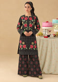 A woman models a black three-piece traditional outfit with vibrant pink and red floral embroidery and green leaf accents, paired with wide-leg pants. She stands in front of a beige studio backdrop with a decorated table in the background.