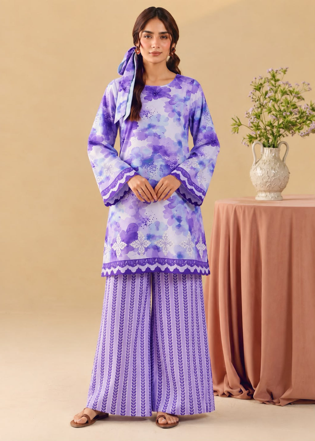 A woman models a traditional Indian outfit consisting of a purple and blue floral printed kurta with wide flared sleeves, paired with matching wide-leg trousers featuring a vertical pattern, set against a warm beige background with a decorative table and vase.