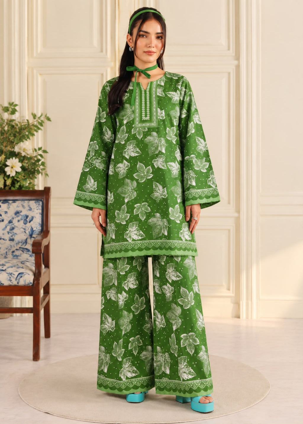A woman modeling a green two-piece kurta and trouser set with a white floral print, standing in a cream-colored room with a patterned chair and a round rug.