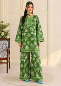 A woman modeling a green two-piece kurta and trouser set with a white floral print, standing in a cream-colored room with a patterned chair and a round rug.