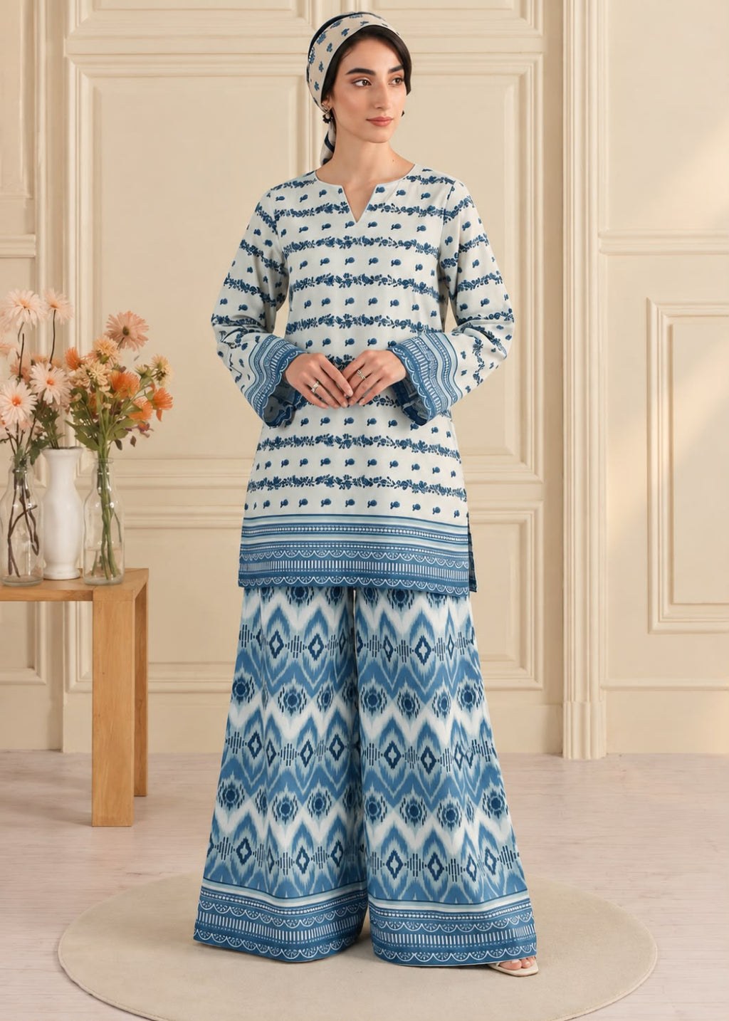 A woman models a two-piece traditional outfit with a white base and blue geometric patterns, consisting of a long-sleeve kurta and wide-leg pants, in an indoor setting with a wooden side table and flowers.