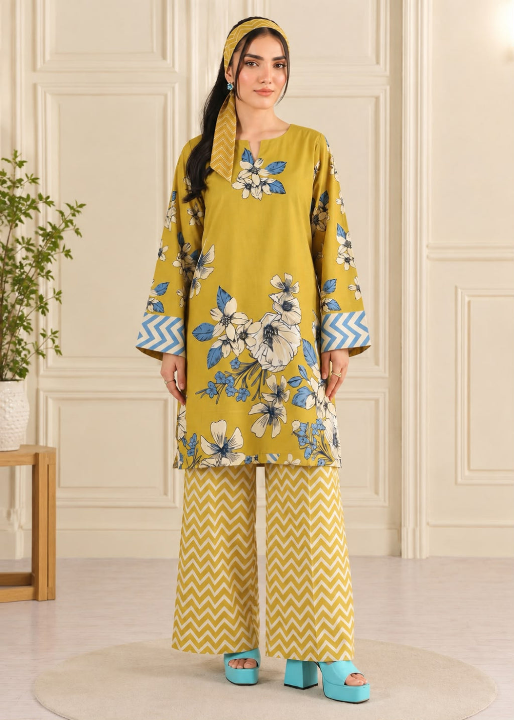 A woman modeling a mustard yellow three-piece kurta set with blue and white floral prints on the tunic, blue chevron sleeve accents, matching wide-leg pants with a yellow and white chevron pattern, a matching headscarf, and turquoise platform sandals.
