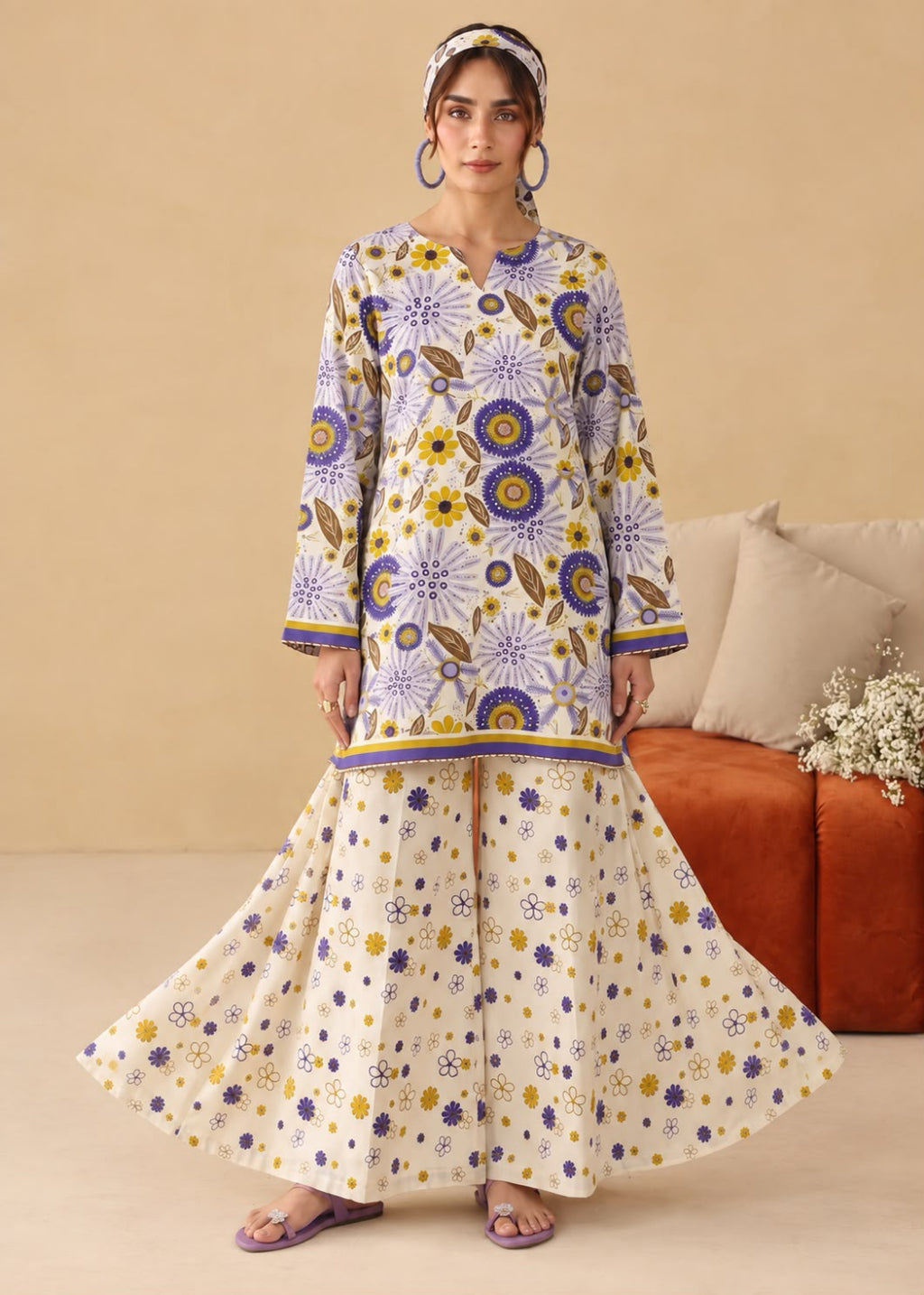 A woman models a traditional long kurta and flared sharara pants set in cream with a vibrant purple and yellow floral print, styled with a matching headband and earrings in a warm beige room.