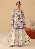 A woman models a traditional long kurta and flared sharara pants set in cream with a vibrant purple and yellow floral print, styled with a matching headband and earrings in a warm beige room.