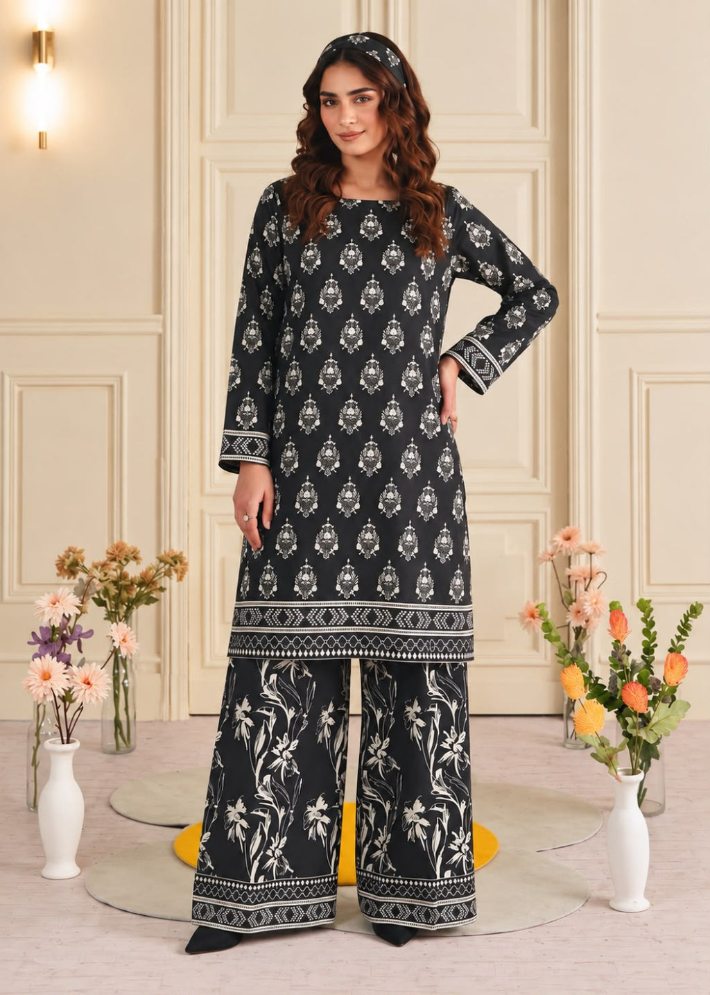 A woman models a black three-piece salwar kameez suit with white floral and geometric patterns, standing in a decorated room with floral arrangements.