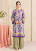 A woman models a traditional three-piece salwar kameez set featuring a purple and yellow floral print kurta with green and yellow striped palazzo pants, set against an elegant cream paneled wall background.
