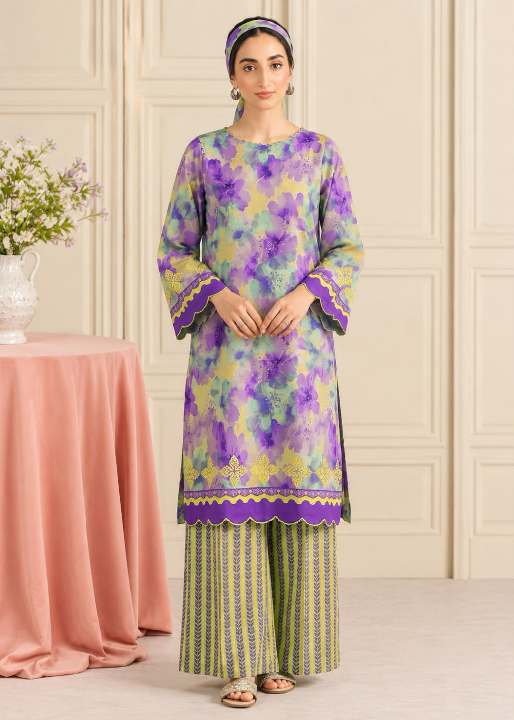A woman models a traditional three-piece salwar kameez set featuring a purple and yellow floral print kurta with green and yellow striped palazzo pants, set against an elegant cream paneled wall background.