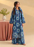 A woman models a blue and white tie-dye long kurta with matching wide-leg pants in a studio setting with a beige background and a pink draped table with a vase of flowers.