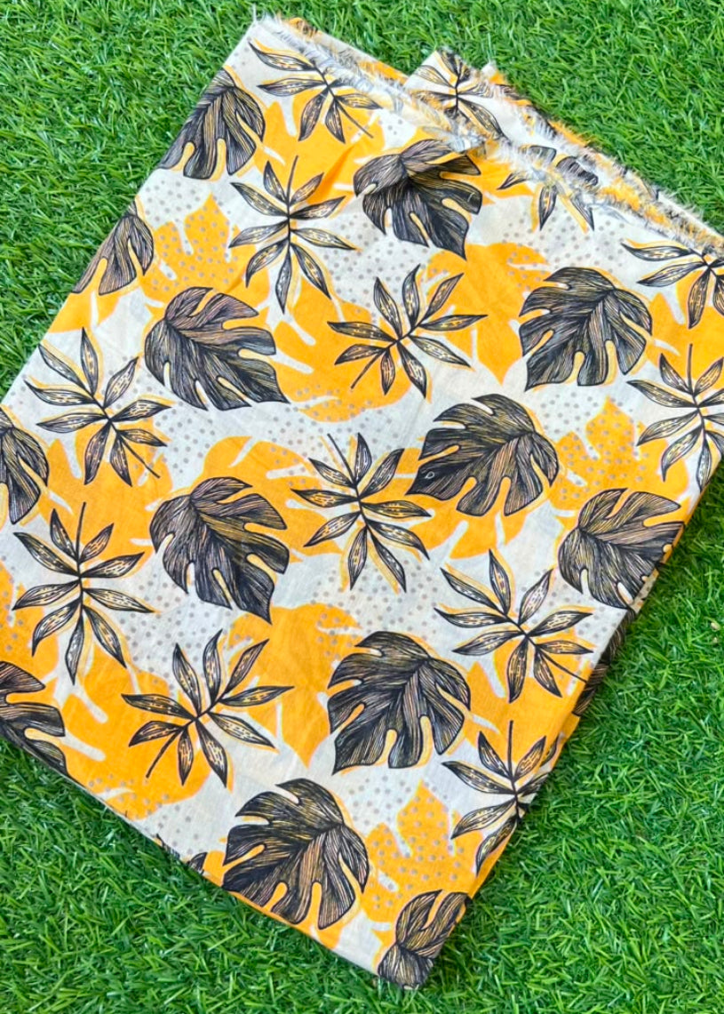 A square fabric item, likely a napkin or small cloth square, featuring a tropical leaf pattern in yellow, dark brown, and white, laid flat on green grass.