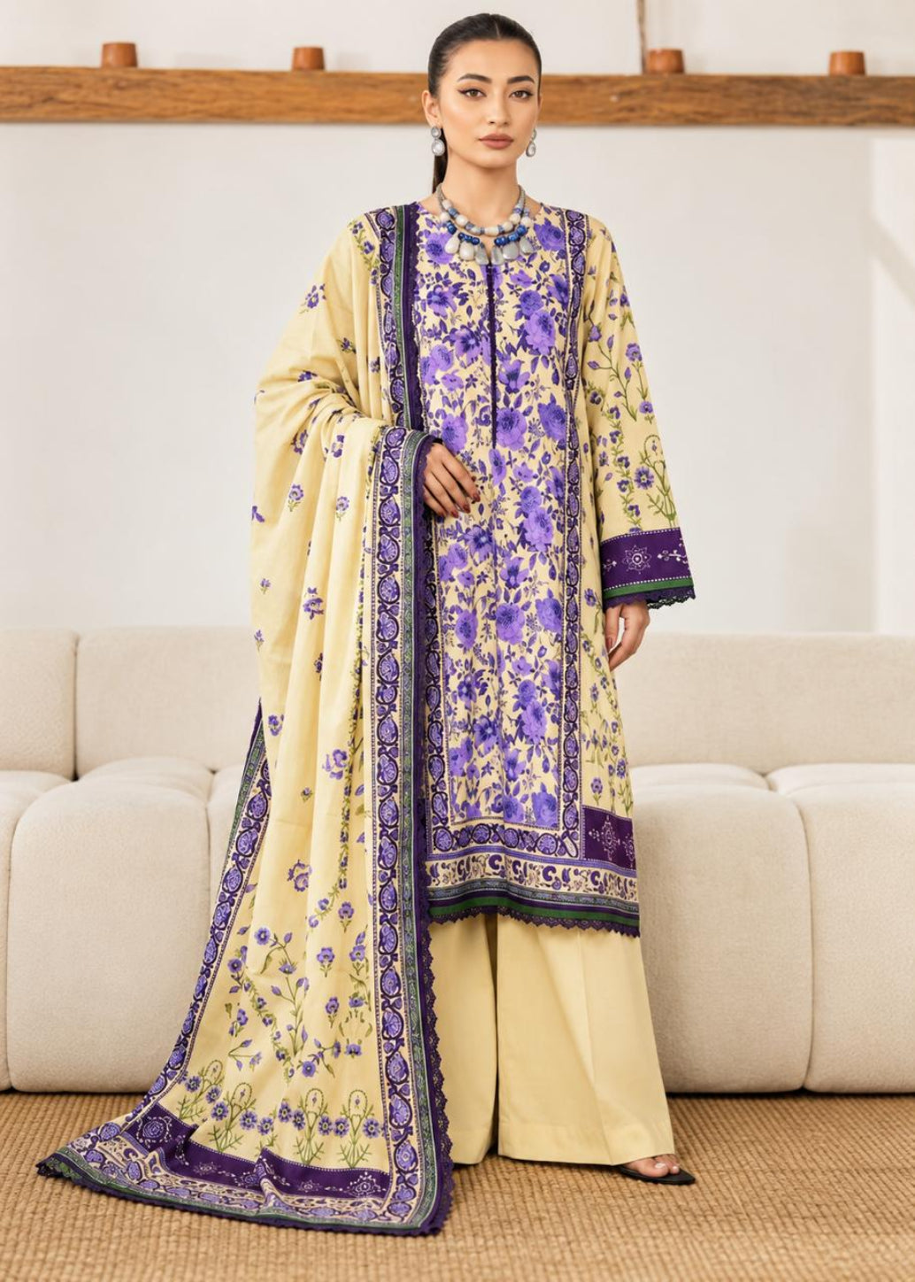 A woman models a traditional South Asian three-piece salwar kameez suit set. The set features a cream-colored kurta and wide-leg trousers with purple floral embroidery and borders, paired with a matching cream dupatta that has a purple border and floral print.