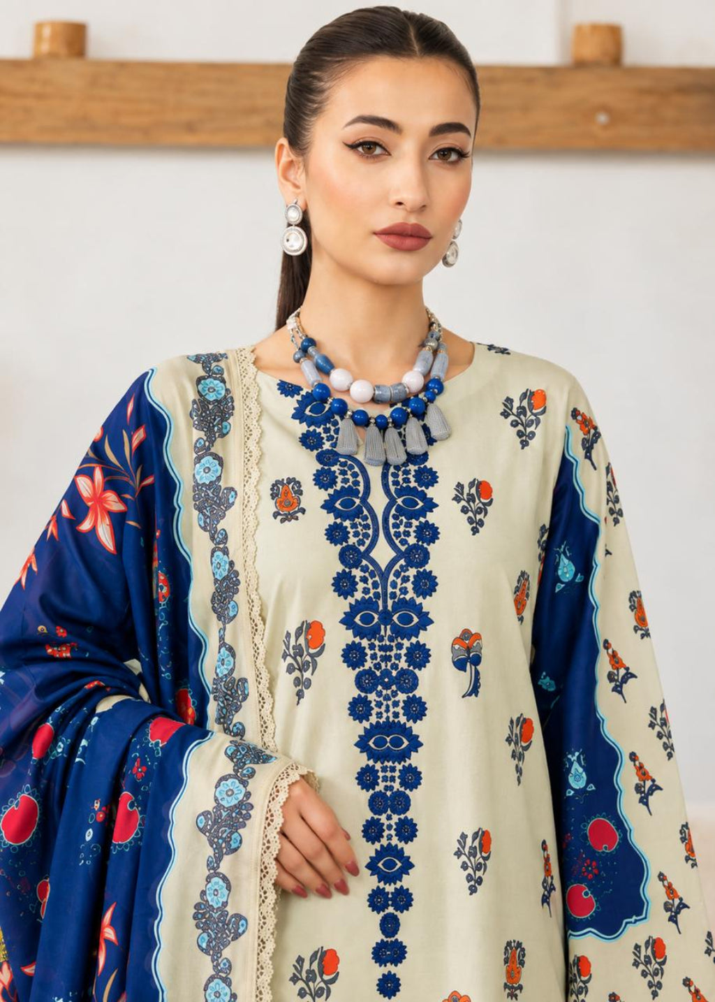 A woman modeling a traditional South Asian salwar kameez outfit with a cream-colored kurta featuring blue floral embroidery down the front, blue patterned sleeves, and a navy blue dupatta with red and blue floral motifs.