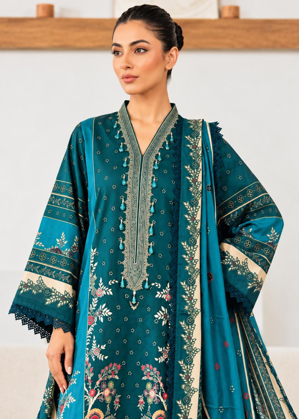 A woman modeling a teal green ethnic three-piece suit with beige and multicolor floral prints, featuring a long kurta, matching trousers, and a dupatta.