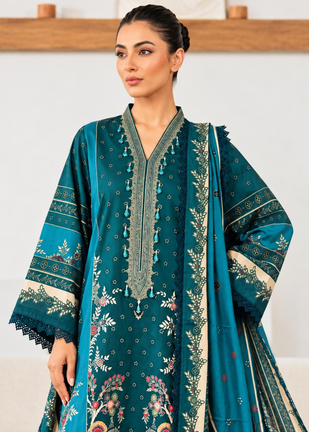A woman modeling a teal green ethnic three-piece suit with beige and multicolor floral prints, featuring a long kurta, matching trousers, and a dupatta.