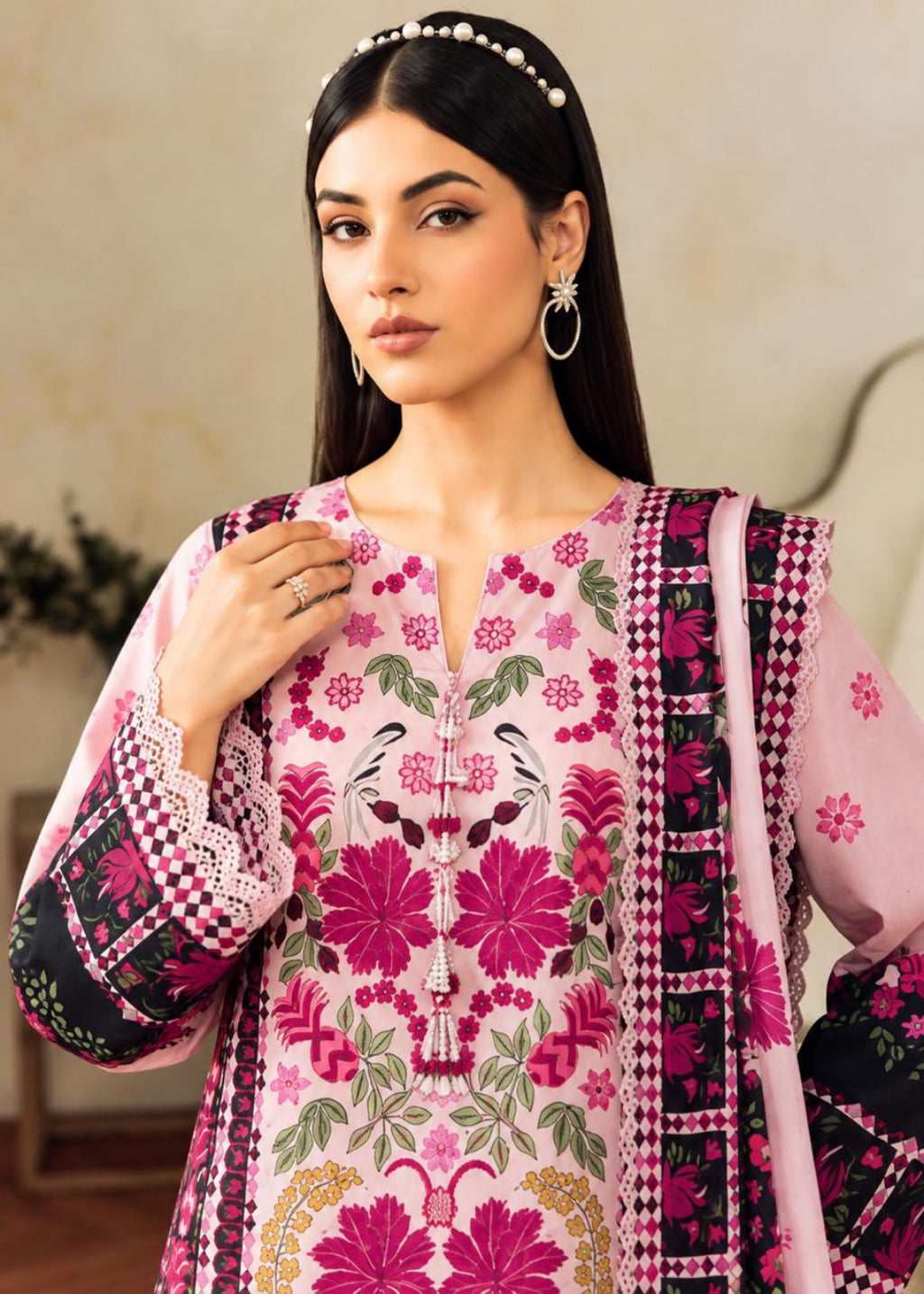 A woman modeling a pink embroidered traditional outfit with floral patterns, black and pink patterned borders, and a matching dupatta, set against a softly blurred indoor background.