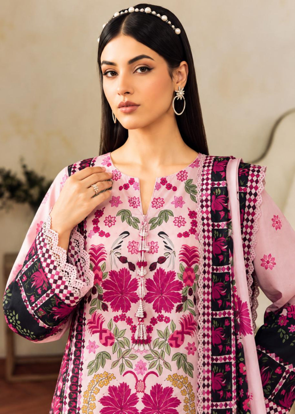 A woman modeling a pink embroidered traditional outfit with floral patterns, black and pink patterned borders, and a matching dupatta, set against a softly blurred indoor background.
