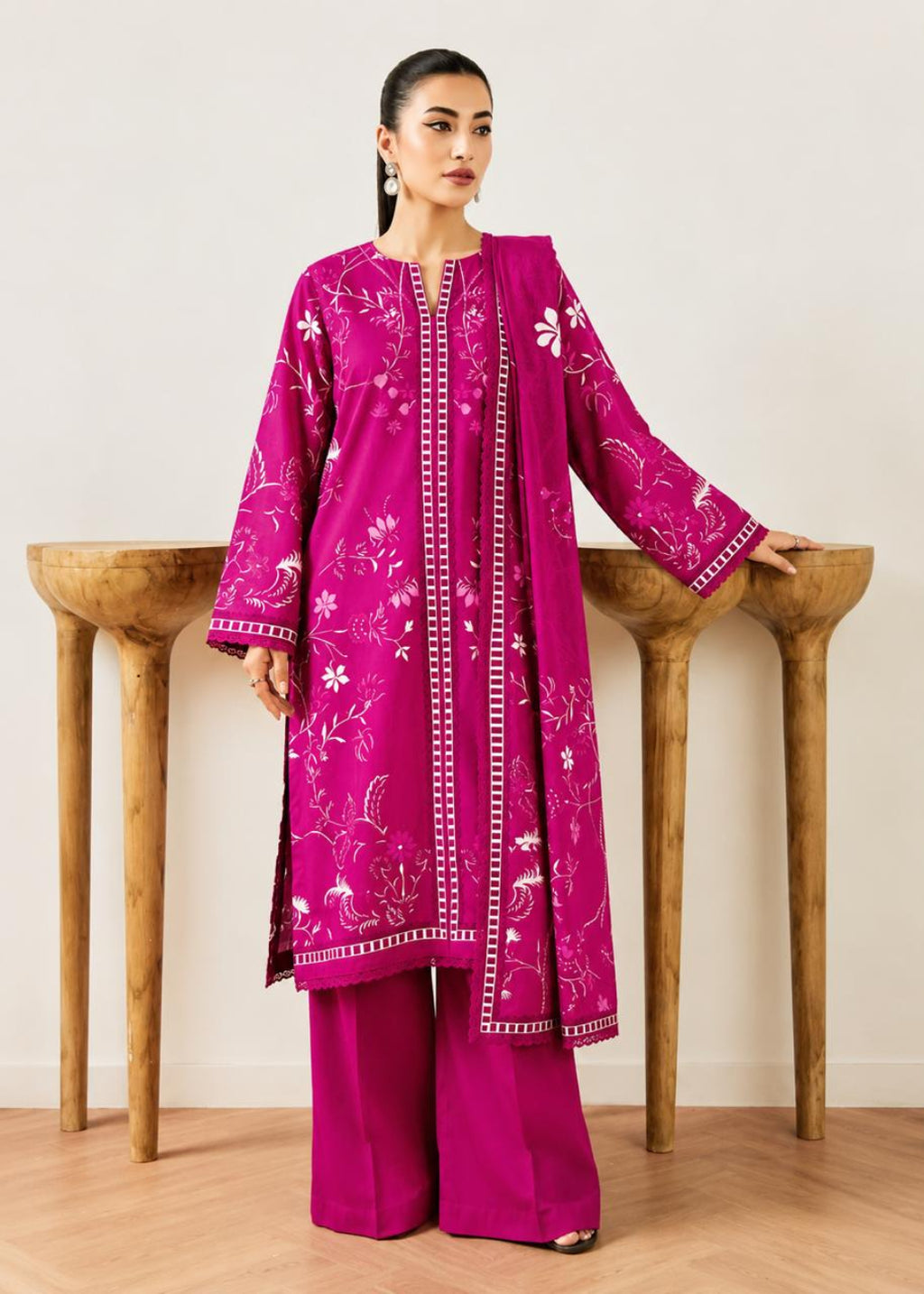 A woman models a magenta pink traditional South Asian outfit, featuring a long kurta with white floral embroidery, matching wide-leg trousers, and a coordinating dupatta, posed in front of a minimalist wooden wall display.