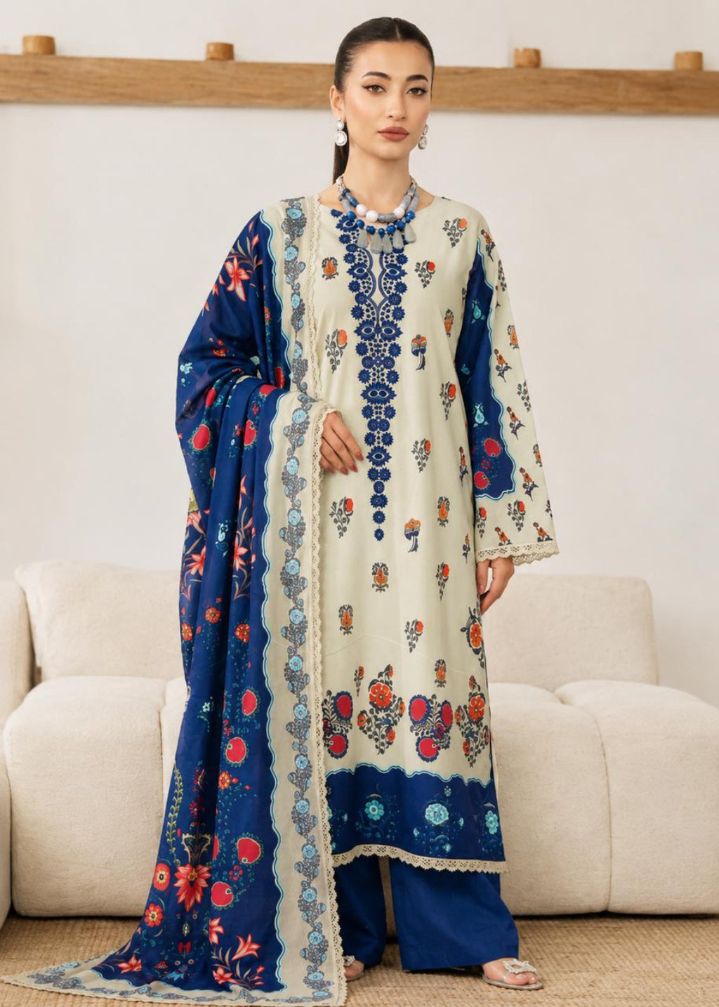 A woman models a traditional three-piece salwar kameez suit set. The set features a cream-colored kurta with blue and red floral embroidery, matching blue trousers, and a navy blue dupatta with intricate floral patterns and borders.