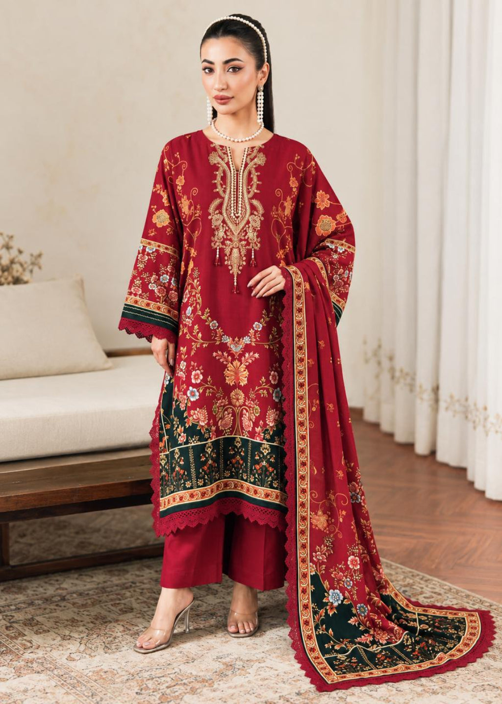 A woman models a traditional three-piece salwar kameez suit in deep red, featuring intricate gold and green floral embroidery, paired with matching red trousers and a long dupatta with a decorative border.