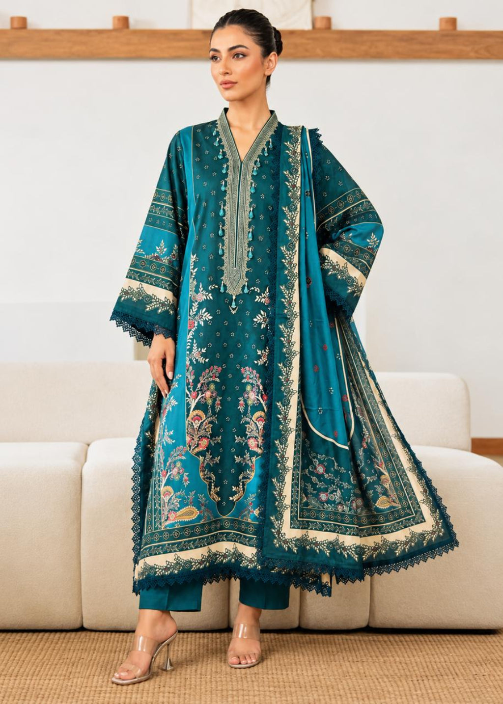 A woman models a traditional teal green three-piece salwar kameez suit with intricate gold and beige embroidery, standing in front of a light-colored sofa.