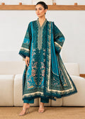 A woman models a traditional teal green three-piece salwar kameez suit with intricate gold and beige embroidery, standing in front of a light-colored sofa.