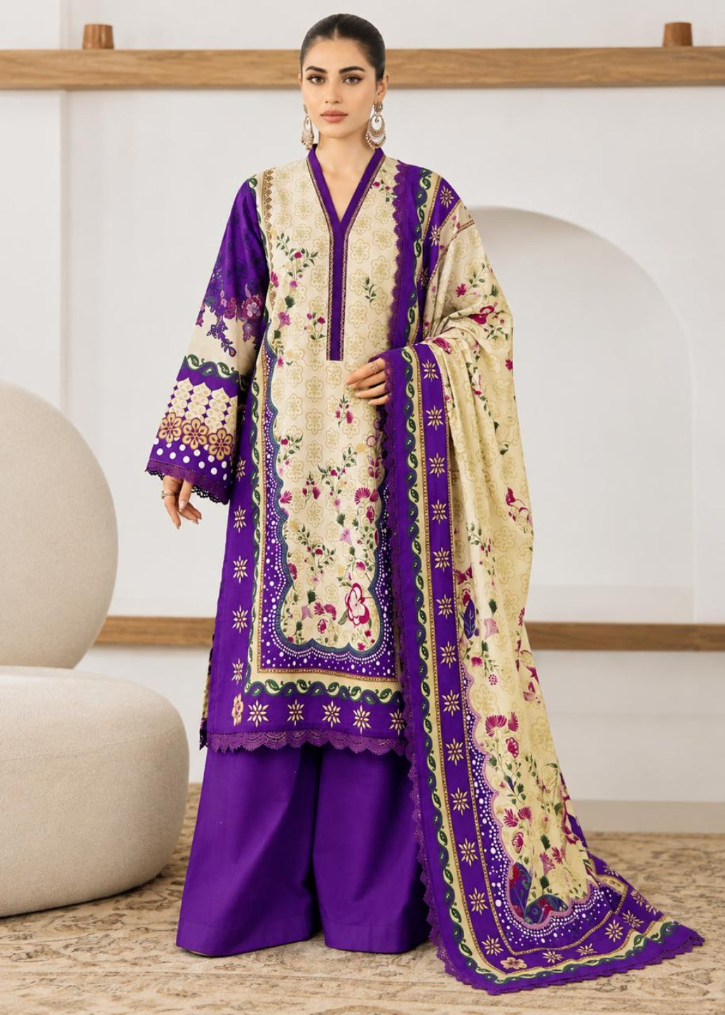 A woman models a traditional South Asian three-piece salwar kameez suit. The set features a deep purple kurta and matching wide-leg trousers, paired with a beige kameez jacket and dupatta heavily decorated with purple borders and floral embroidery.