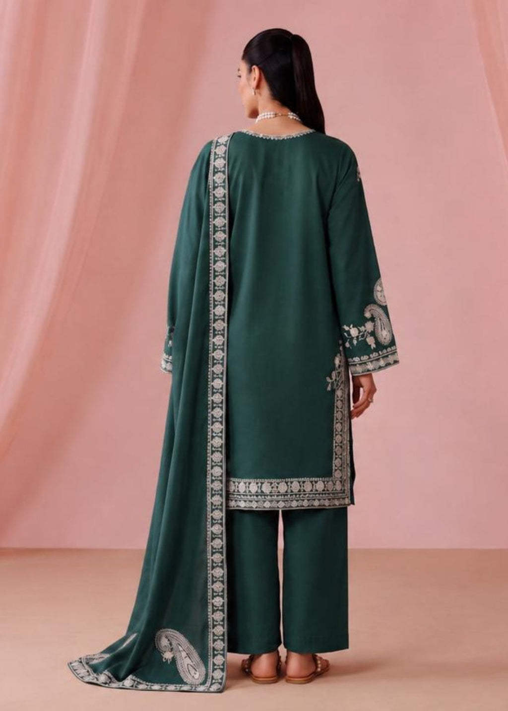 A woman stands with her back to the camera wearing a dark green traditional outfit with silver embroidered borders, consisting of a long kurta, pants, and a dupatta, set against a pink draped backdrop.