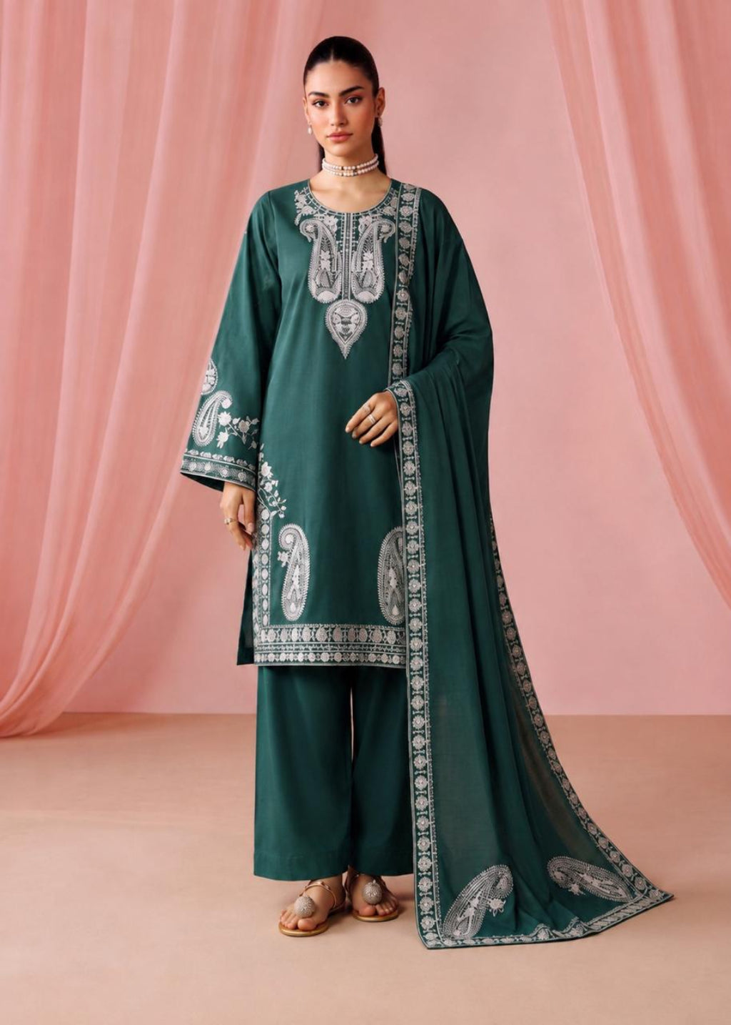 A woman models a dark green traditional outfit with silver embroidery, matching wide-leg pants, and a sheer dupatta draped over a pink draped fabric backdrop.