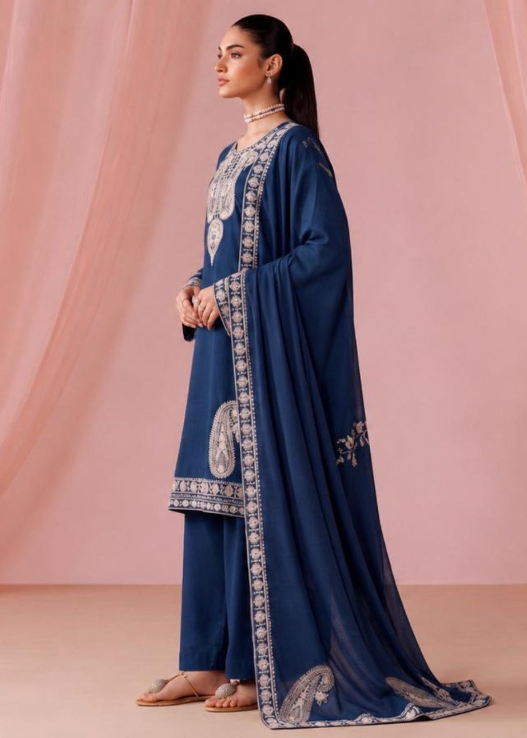 A woman stands in profile wearing a navy blue traditional outfit with ornate silver embroidery and a matching draped scarf, posed against a soft pink draped fabric backdrop.