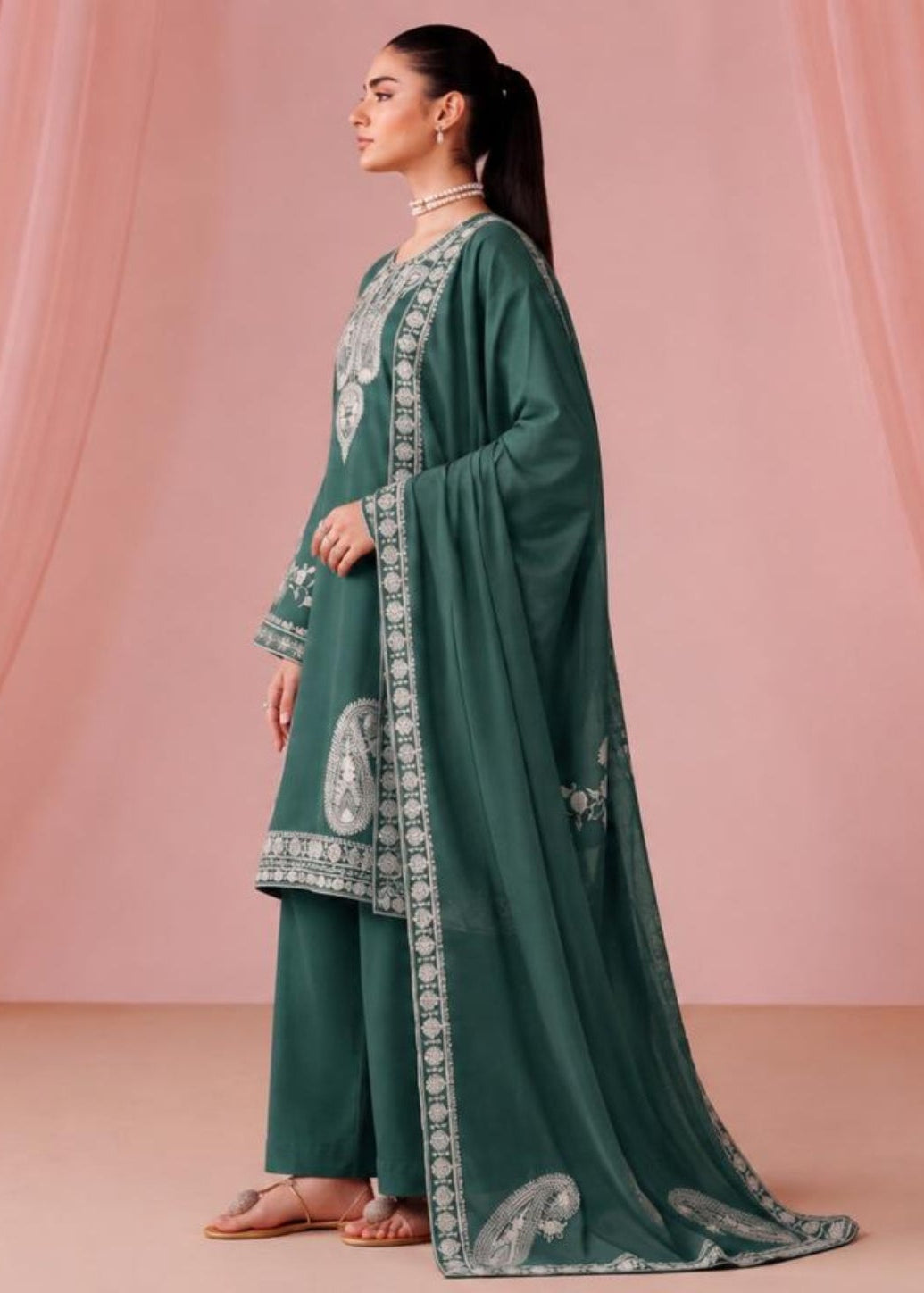 A woman stands in profile wearing a dark teal green traditional outfit with white embroidered borders and matching draped fabric, posed against a soft pink draped backdrop.