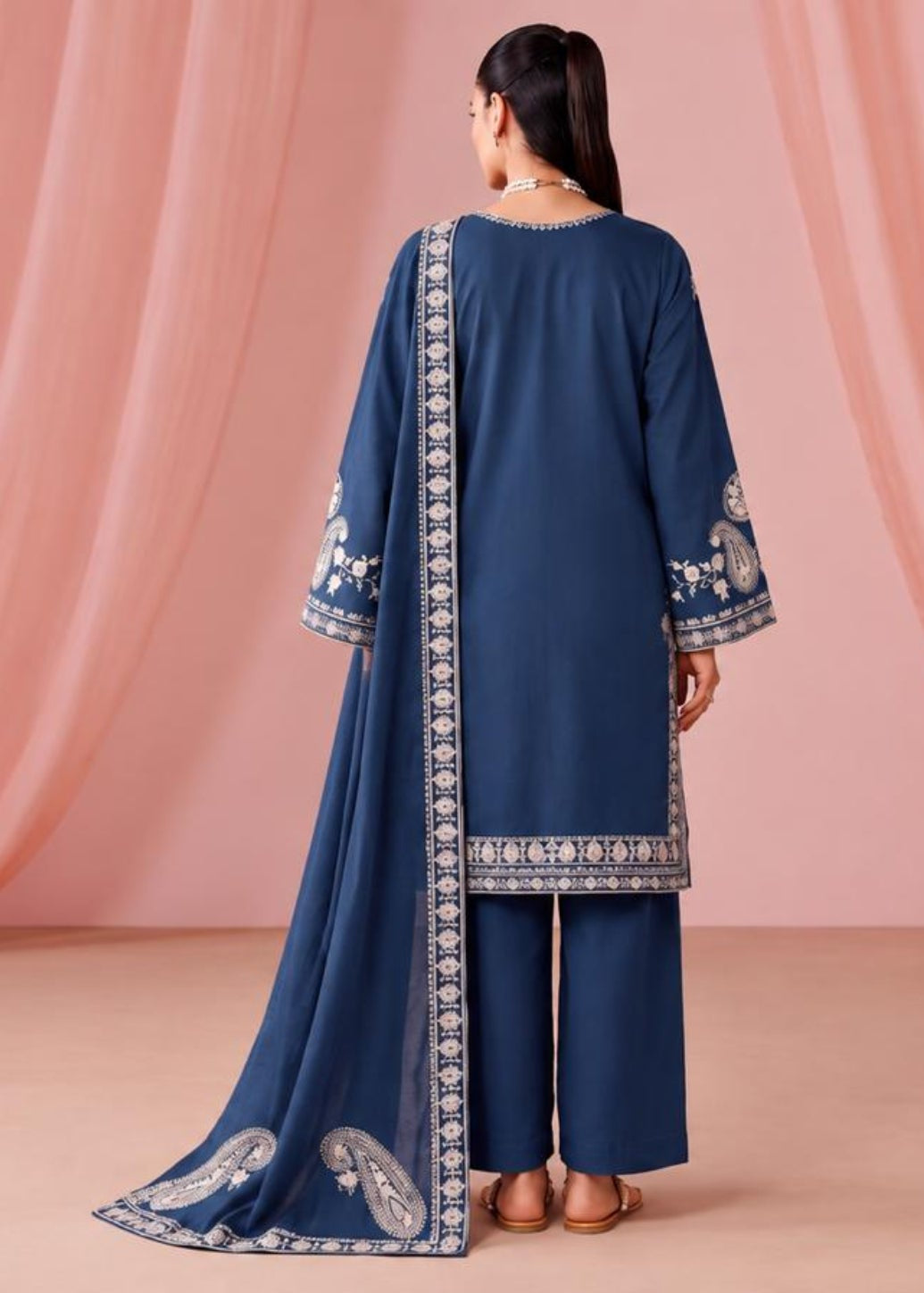 A woman stands with her back to the camera wearing a navy blue traditional outfit with white embroidered borders and matching pants, posed in front of draped pink fabric.