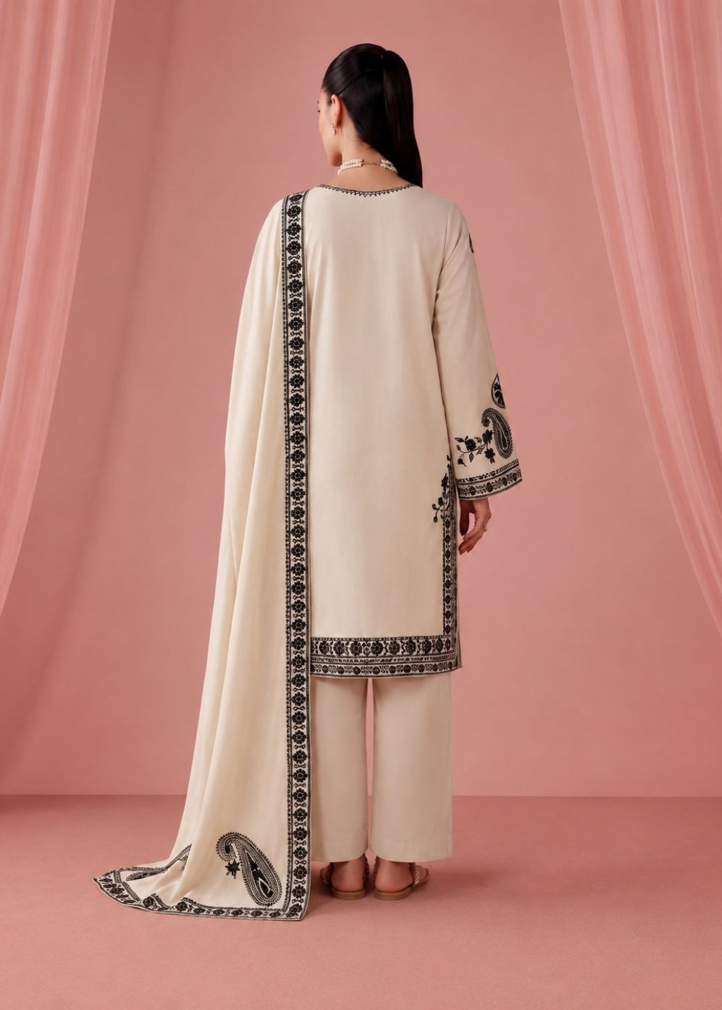 A woman stands with her back to the camera wearing a cream-colored traditional outfit with black embroidered borders and a matching dupatta, set against a pink draped backdrop.