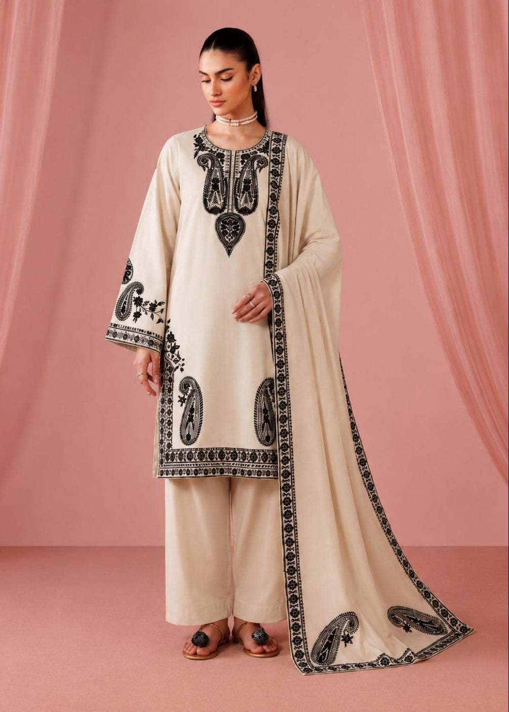 A woman models a traditional Indian outfit consisting of a cream-colored kurta with black embroidery, matching wide-leg trousers, and a coordinating dupatta, set against a pink draped backdrop.