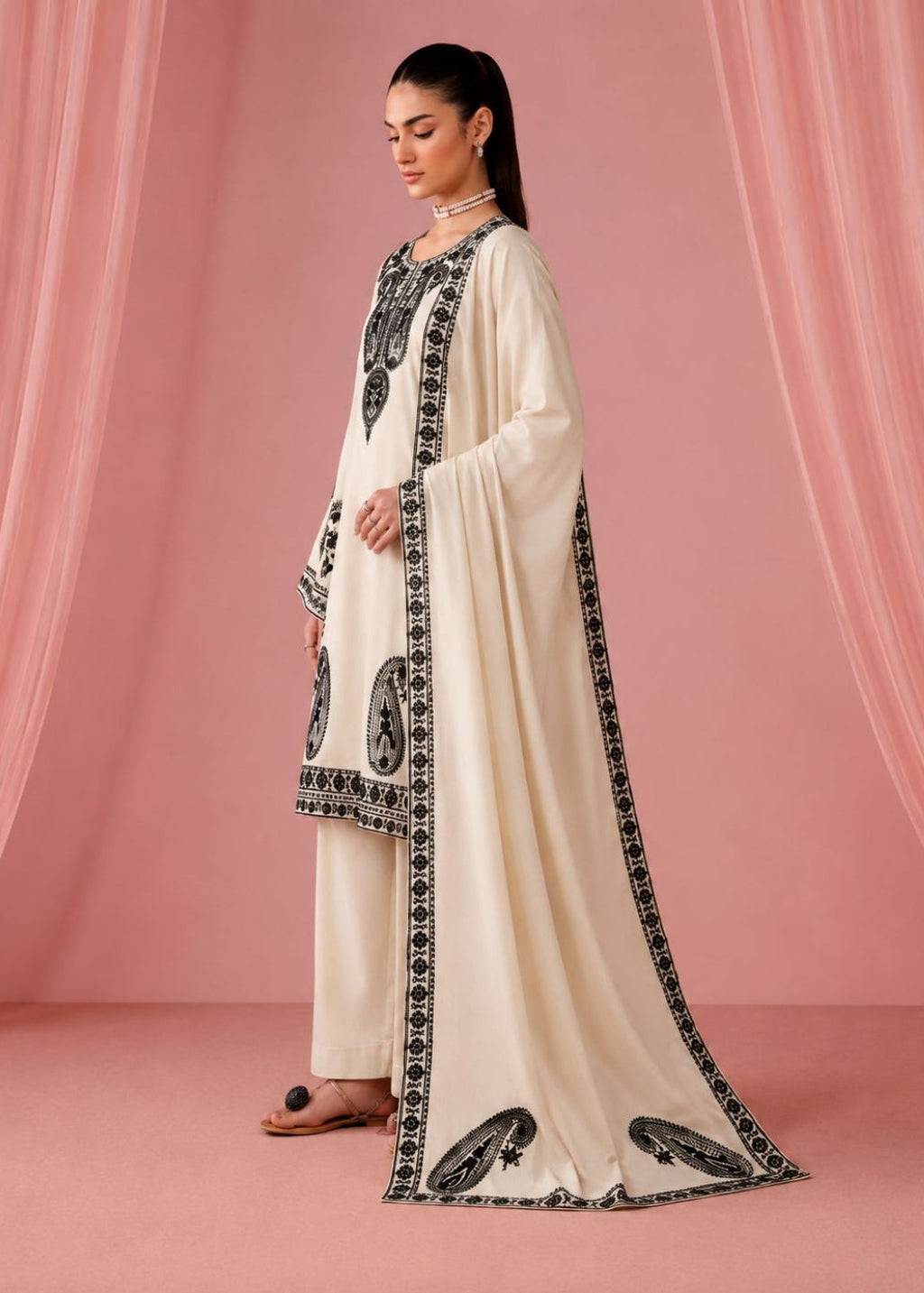 A woman models a traditional cream-colored three-piece outfit with black embroidered details and a matching dupatta, set against a pink draped backdrop.