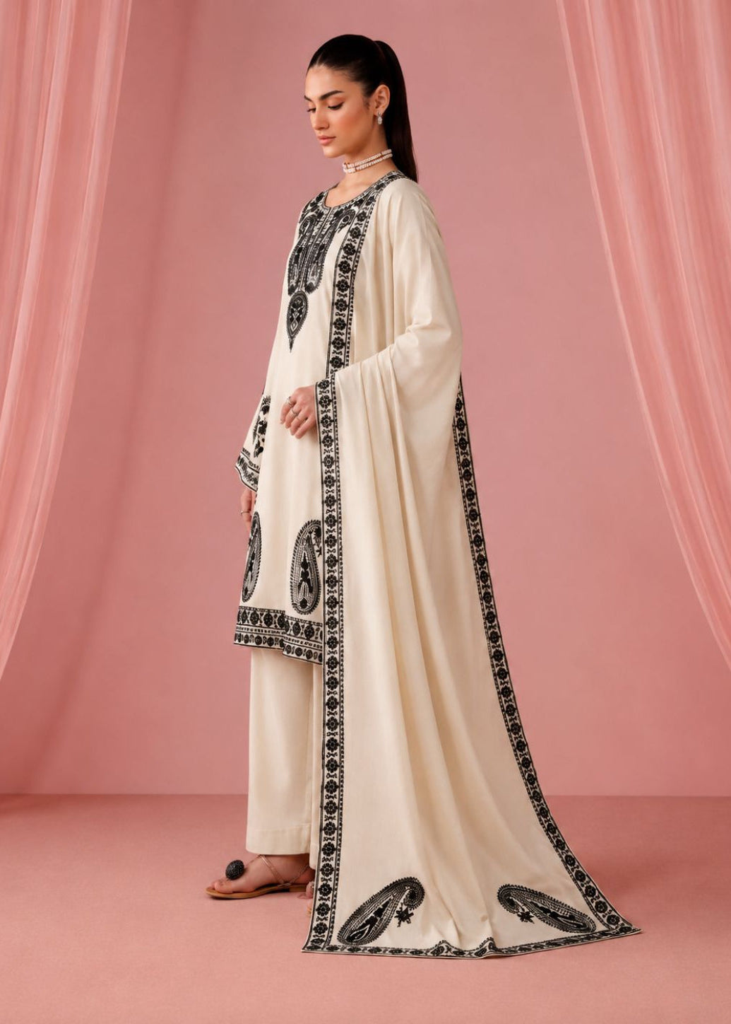 A woman models a traditional cream-colored three-piece outfit with black embroidered details and a matching dupatta, set against a pink draped backdrop.