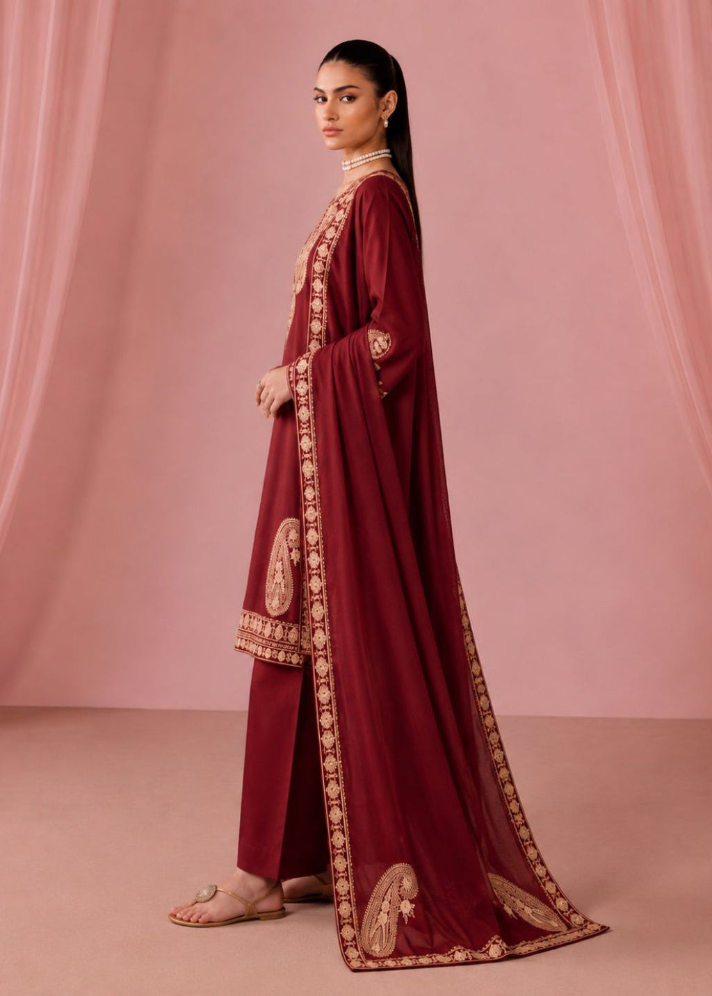 A woman models a deep maroon traditional outfit with gold embroidery, consisting of a long kurta, matching trousers, and a sheer dupatta, set against a soft pink draped backdrop.