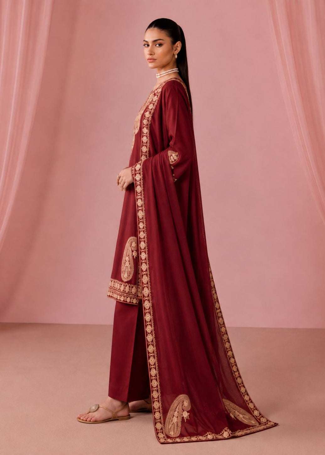 A woman models a deep maroon traditional outfit with gold embroidery, consisting of a long kurta, matching trousers, and a sheer dupatta, set against a soft pink draped backdrop.