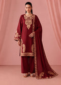 A woman models a traditional Indian salwar kameez suit in deep maroon with intricate gold embroidery, paired with matching maroon trousers and a long dupatta, set against a draped pink backdrop.