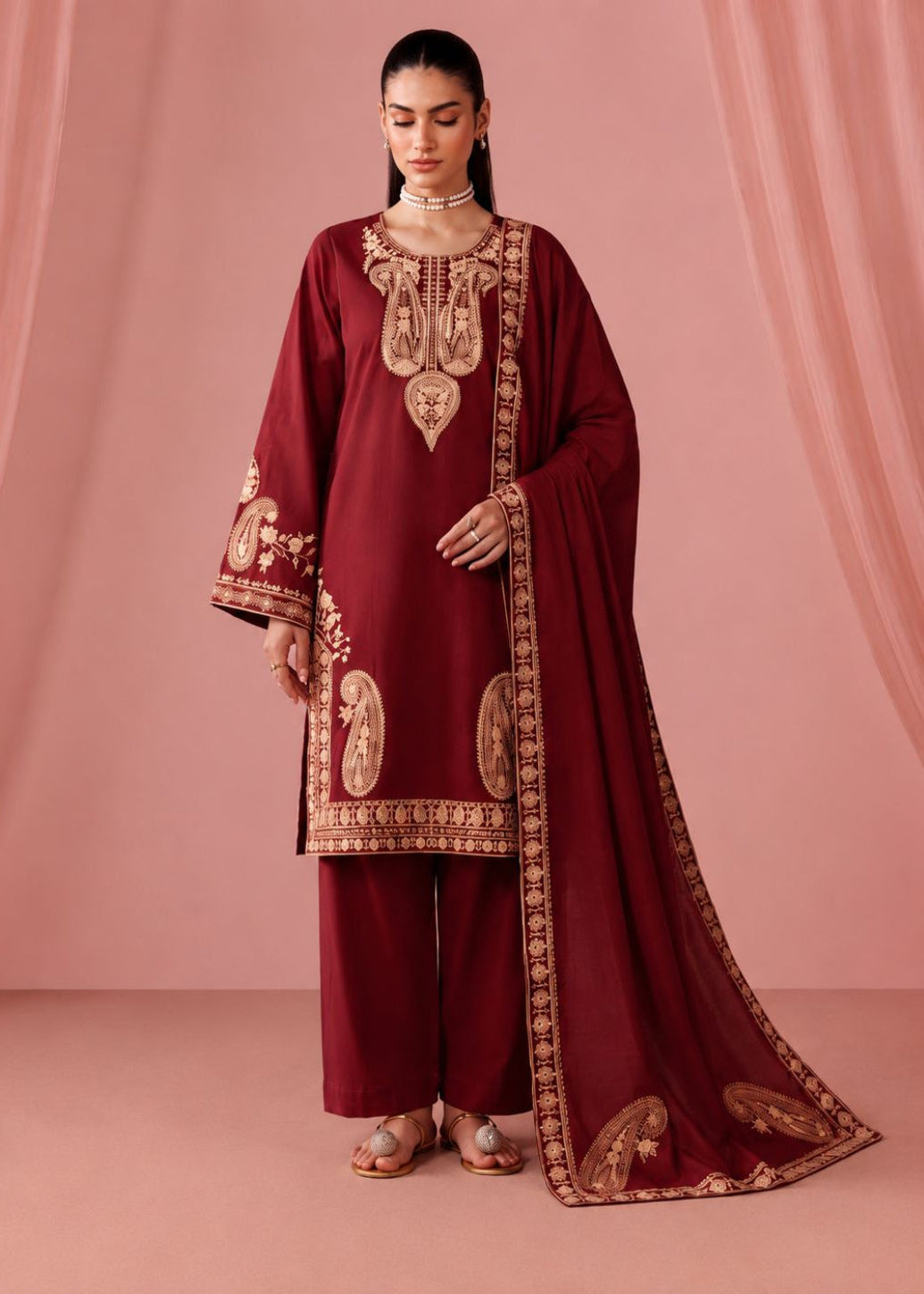 A woman models a traditional Indian salwar kameez suit in deep maroon with intricate gold embroidery, paired with matching maroon trousers and a long dupatta, set against a draped pink backdrop.
