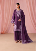 A woman models a deep purple three-piece traditional outfit with matching printed dupatta, featuring a kurta with ornate light purple embroidery, wide-leg trousers, and a draped scarf, set against a soft pink draped backdrop.