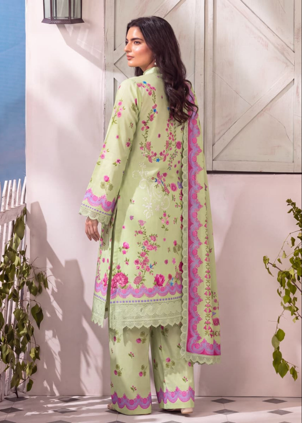 A woman modeling a light green three-piece traditional outfit with pink floral embroidery and purple border details, shown from the back in an outdoor patio setting.