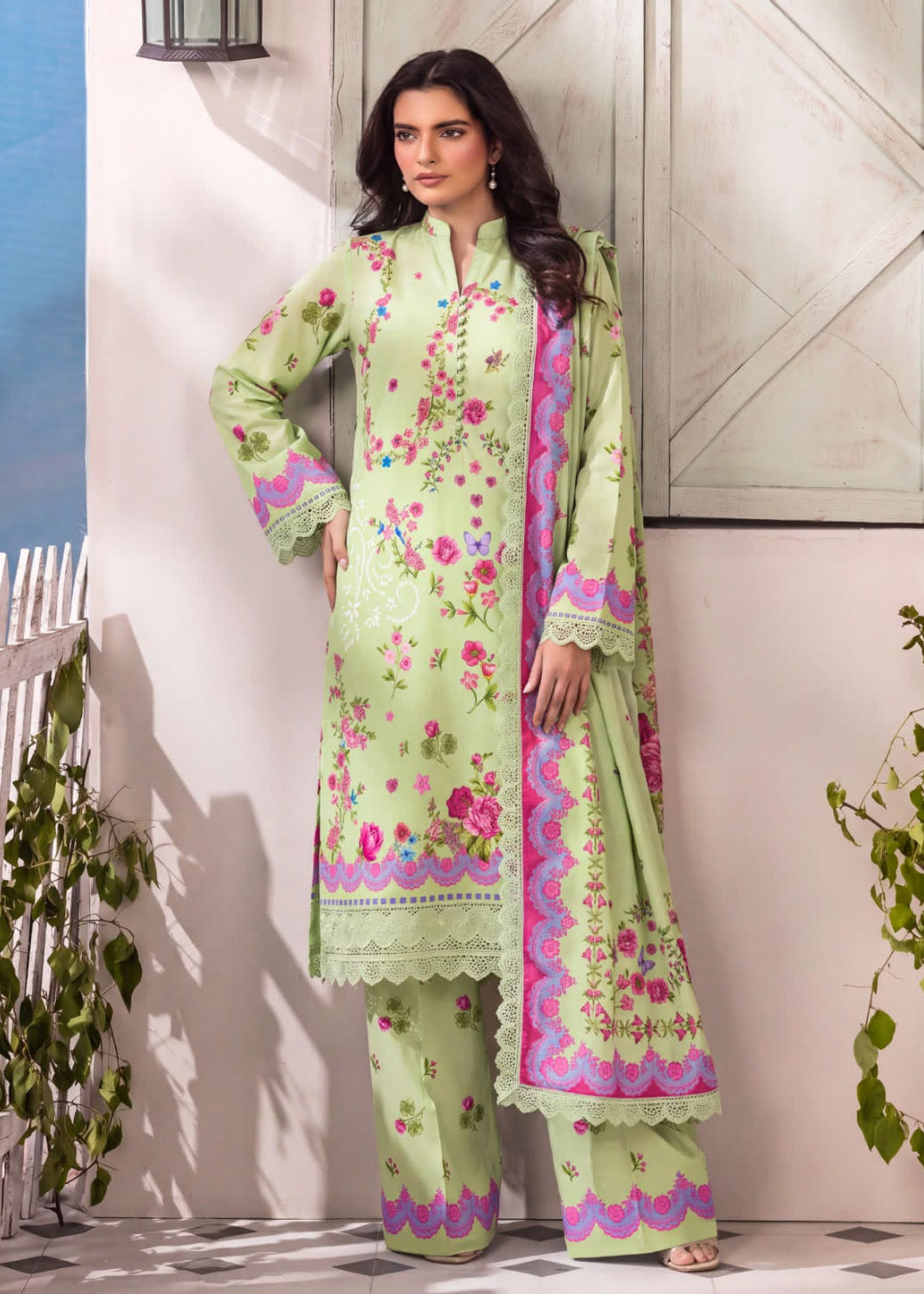 A woman models a light green three-piece traditional outfit with pink and purple floral embroidery and border details, standing against a rustic white wooden door and wall.