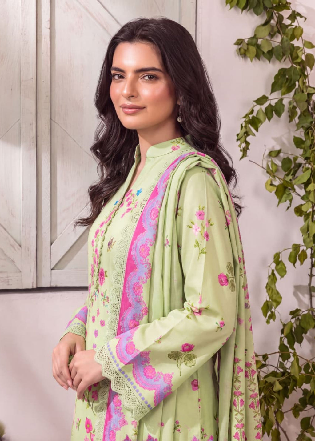 A woman wearing a light green traditional outfit with pink and purple floral embroidery and lace details, posed against a rustic white paneled wall with green foliage on the side.