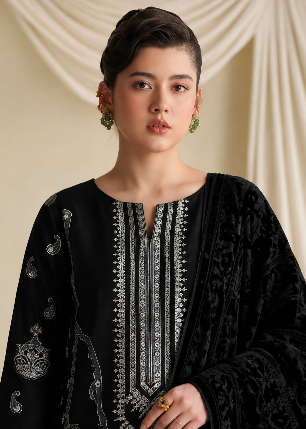 A woman modeling a black traditional outfit with intricate silver embroidery and a matching black velvet dupatta, set against a draped cream fabric background.